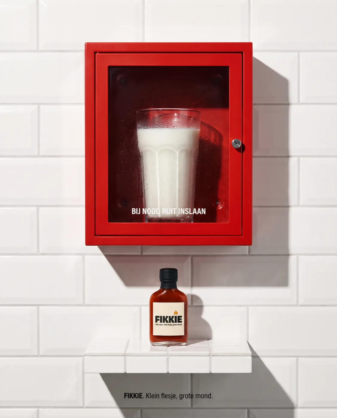 Rood noodkastje met een glas melk achter glas — bij nood ruit inslaan — met het FIKKIE-flesje eronder